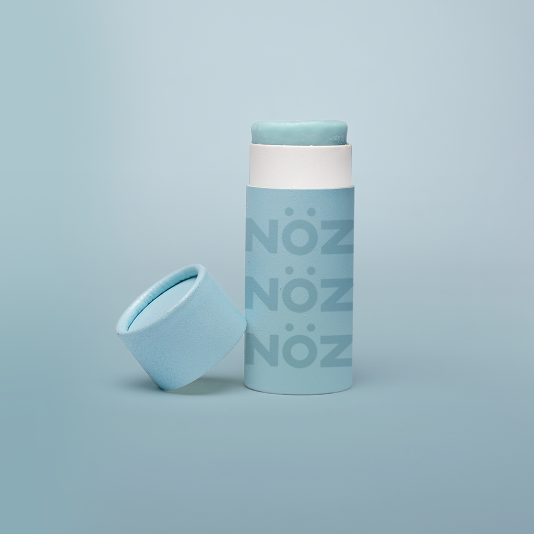 Noz' blue nose sunscreen with the cap open and leaning up against the side of the cylinder sunscreen container, with a light blue background.