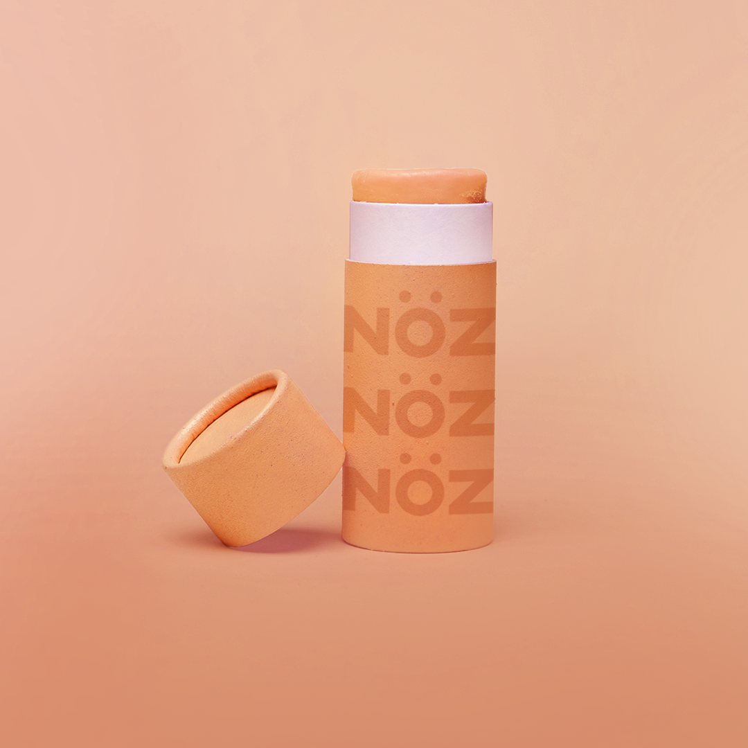 Noz' orange nose sunscreen with the cap open and leaning up against the side of the cylinder sunscreen container, with a light orange background.