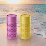 Load image into Gallery viewer, Noz&#39; purple and yellow nose sunscreens on a beach with a beautiful sunset in the background. 
