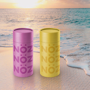 Noz' purple and yellow nose sunscreens on a beach with a beautiful sunset in the background. 